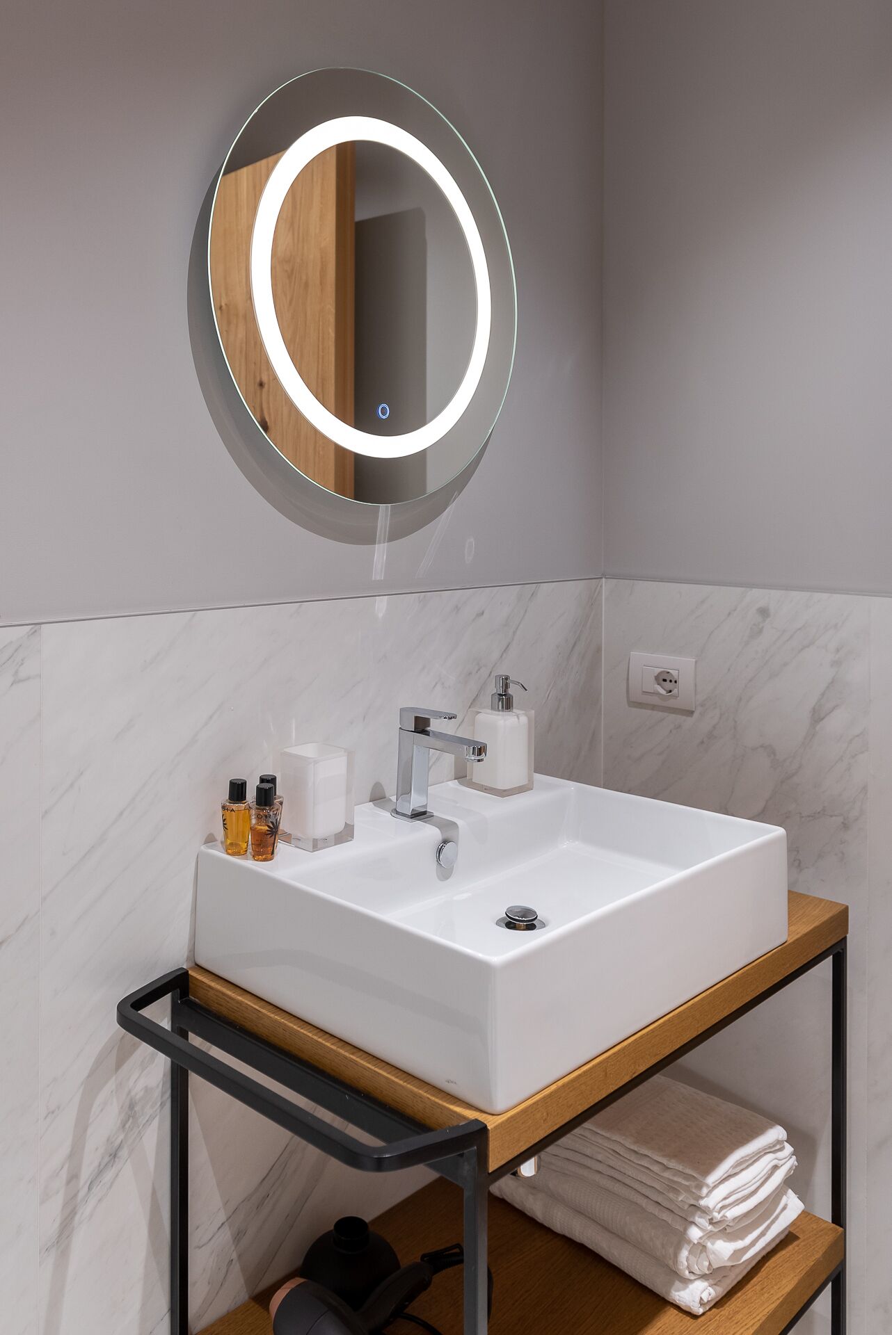 Un baño moderno con un lavamanos blanco, un espejo redondo con luz, un estante de madera con toallas y una pared de mármol.