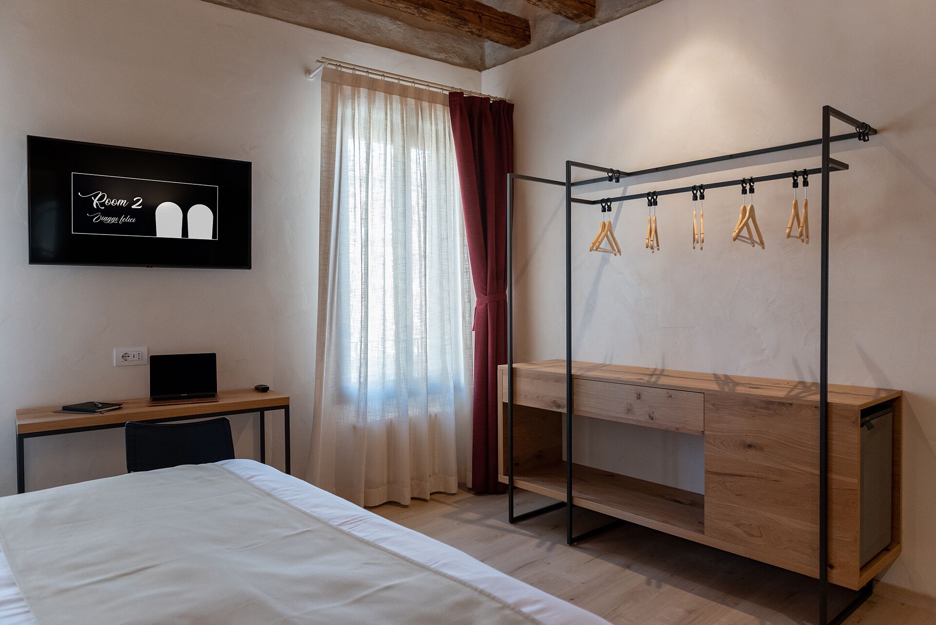 Une chambre d’hôtel avec un lit, un bureau équipé d’un ordinateur, un téléviseur affichant le numéro « 2 », et une armoire en bois avec une tringle pour suspendre les vêtements.