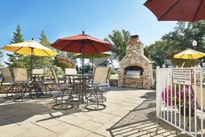 An outdoor patio with tables and chairs, a stone fireplace, and umbrellas. - The Savannah: 2BR + Loft Townhome Sleeps 8 | Pool, Hot Tub, Free Golf, Near Lake Michigan (Holland)
