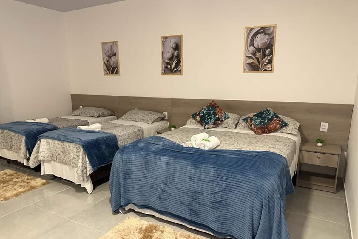 Habitación de hotel con tres camas, cada una con un edredón de estampado diferente. Se observan tres cuadros florales colgados en la pared, una mesita de noche con una planta y una alfombra mullida en el suelo.