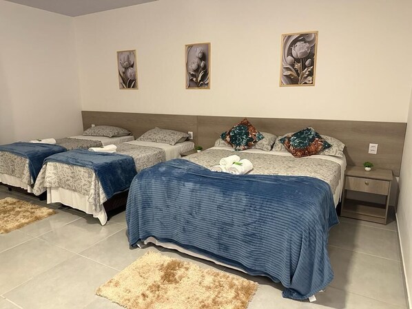 Habitación de hotel con tres camas, cada una con un edredón de estampado diferente. Se observan tres cuadros florales colgados en la pared, una mesita de noche con una planta y una alfombra mullida en el suelo.