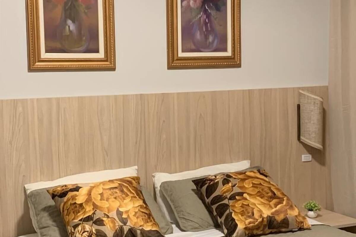 Habitación de hotel con una cama, dos cuadros enmarcados con motivos florales, cabecera de madera y una pequeña planta en maceta.