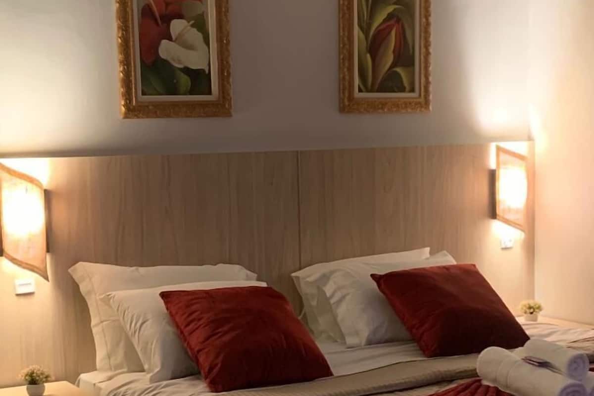 Habitación de hotel con cabecera de madera, dos almohadas rojas, una manta roja y dos cuadros florales enmarcados en la pared.