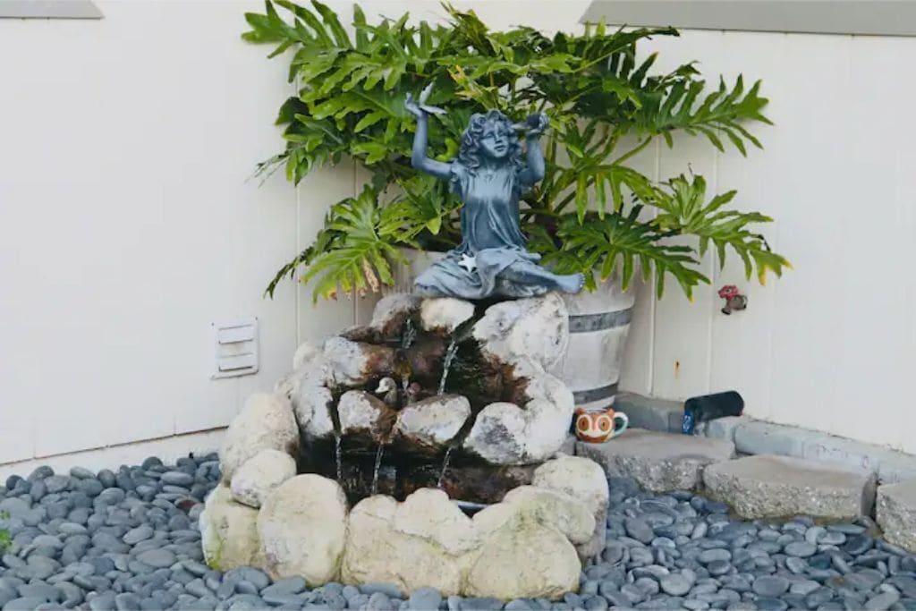 A statue of a woman on a rock with a water feature in front of a white wall and green foliage.