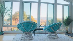 Quarto com janelas amplas, duas cadeiras de vime, uma pequena mesa e um vaso de planta.