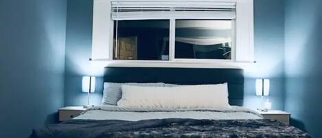 A bedroom with a bed, two bedside tables, a window with blinds, and wall-mounted lamps.