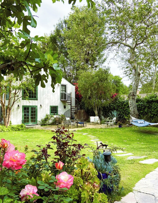 Una casa blanca con puerta y ventanas verdes, rodeada de un jardín cuidado con rosales rosados y una hamaca.