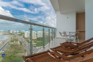 A balcony with wooden lounge chairs, a glass railing, and a view of the beach and cityscape. - Apartamento frente al mar Cartagena (Cartagena)