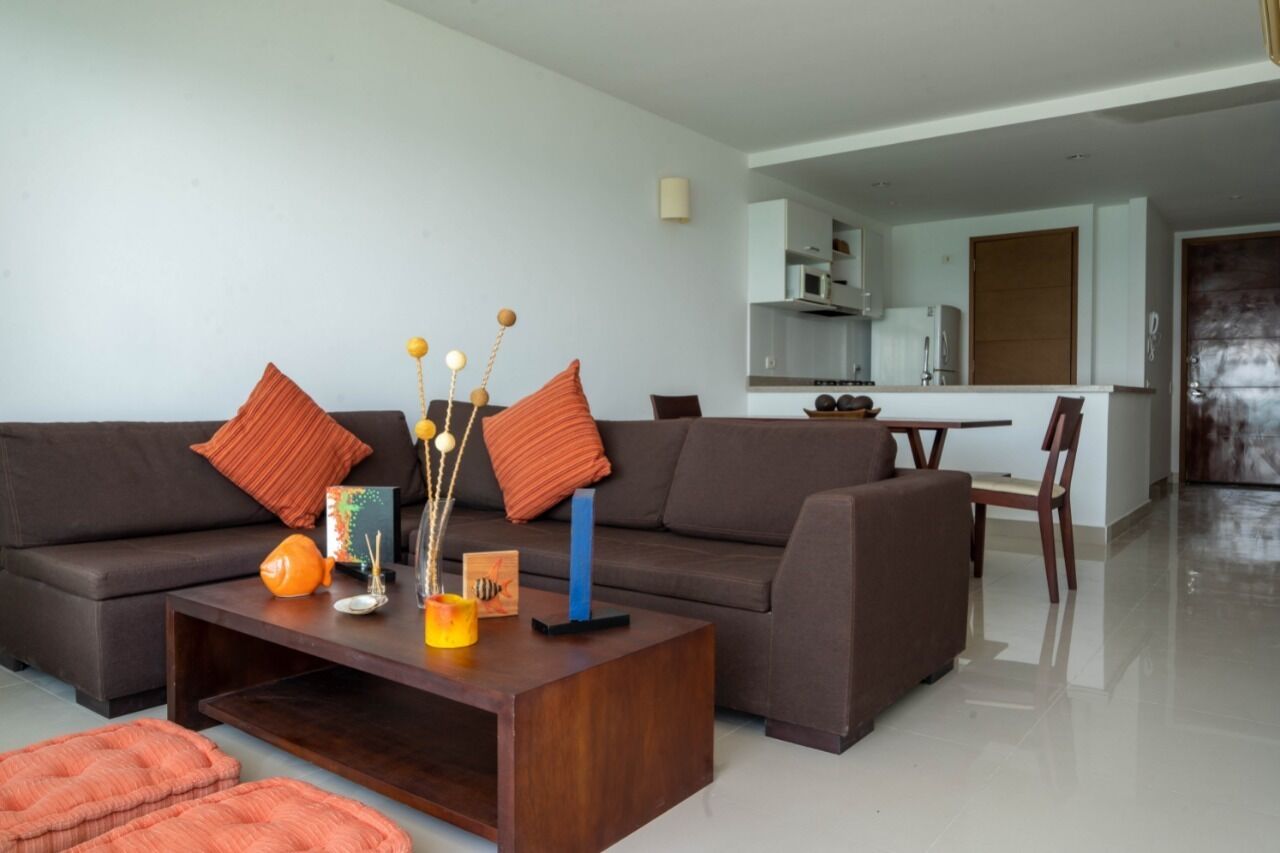 A modern living room with a dark brown sofa, orange cushions, and a wooden coffee table with decorative items. A kitchen is visible in the background.