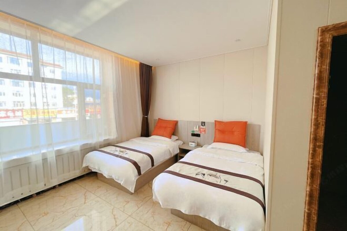 A hotel room with two beds, a window with sheer curtains, a radiator, and a view of buildings outside.