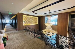 Un hall de hotel con un largo pasillo, un muro decorativo, una escalera con barandilla y una lámpara colgante.