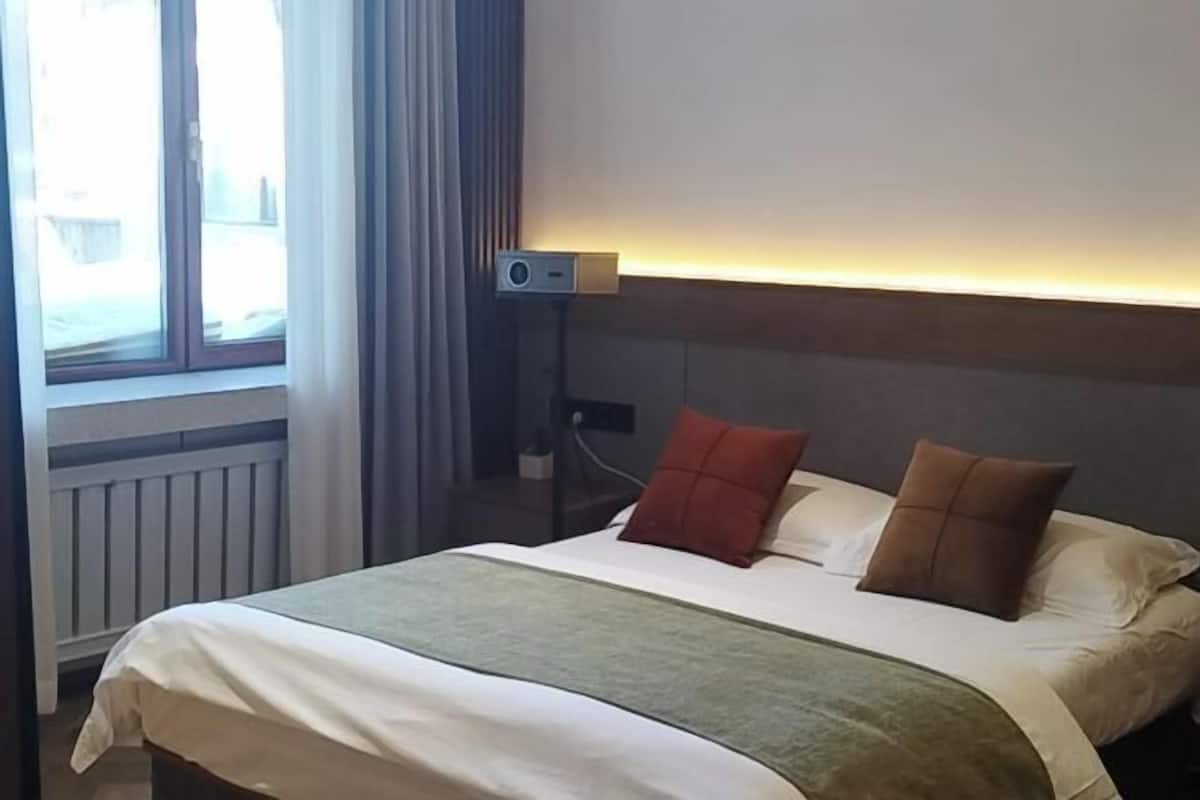 Una habitación de hotel con una cama, una mesita de noche, una lámpara y un ventanal con cortinas.