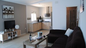 Moderni olohuone, jossa on tummanruskea sohva, lasinen sohvapöytä ja televisio puukehikkoisessa telineessä. Taustalla näkyy keittiö, jossa on puukehikoiset kaapit ja ruostumattomasta teräksestä valmistettu liesituuletin.