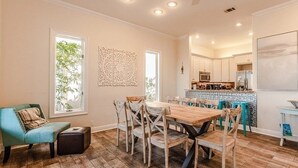 A spacious dining area with a wooden table and chairs, a kitchen in the background, and a window with a view of greenery. - Villa De Palmas 5 Bedroom Home by RedAwning (Destin)