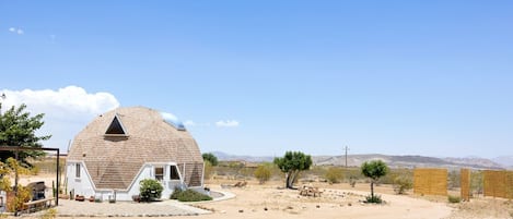A dome-shaped house with a small attached structure, surrounded by a dry landscape with sparse vegetation and distant mountains.