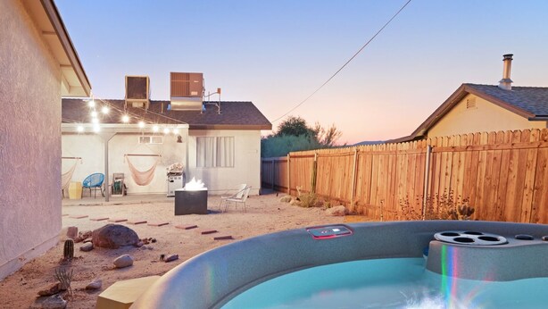 A backyard with a hot tub, a wooden fence, and a house with a chimney. - The Rockhopper - Hot Tub, Fire Pit & Walk To Downtown Jt! 4 Bedroom Home by RedAwning (Joshua Tree)