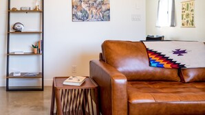 A brown leather sofa with a colorful throw pillow, a wooden side table with a book, a wooden bookshelf with decorative items, and a painting on the wall. - The Rockhopper - Hot Tub, Fire Pit & Walk To Downtown Jt! 4 Bedroom Home by RedAwning (Joshua Tree)
