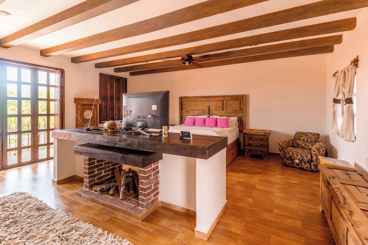 Amplia habitación con techo de madera, chimenea, cama con almohadas rosas, televisor y sillón estampado.