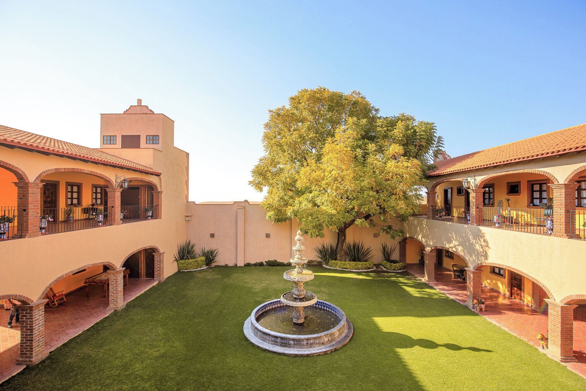 Un patio central con una fuente, rodeado de edificios con paseos arqueados y un árbol.