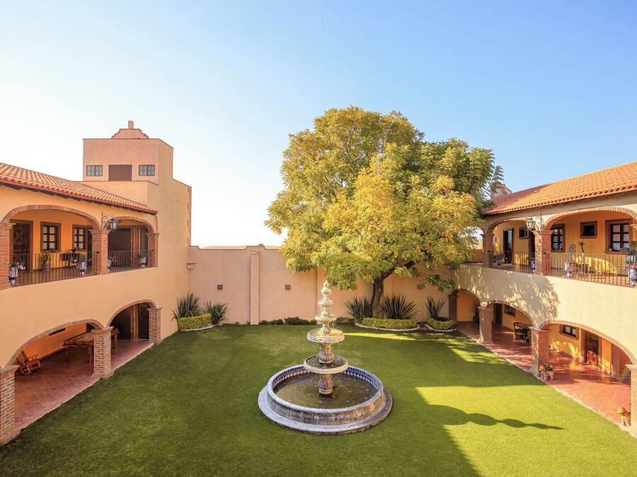 A courtyard with a central fountain, surrounded by buildings with arched walkways and a tree.