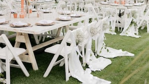 Un espace extérieur aménagé pour les repas, avec des tables et des chaises blanches, des parasols beiges et de la verdure en arrière-plan.