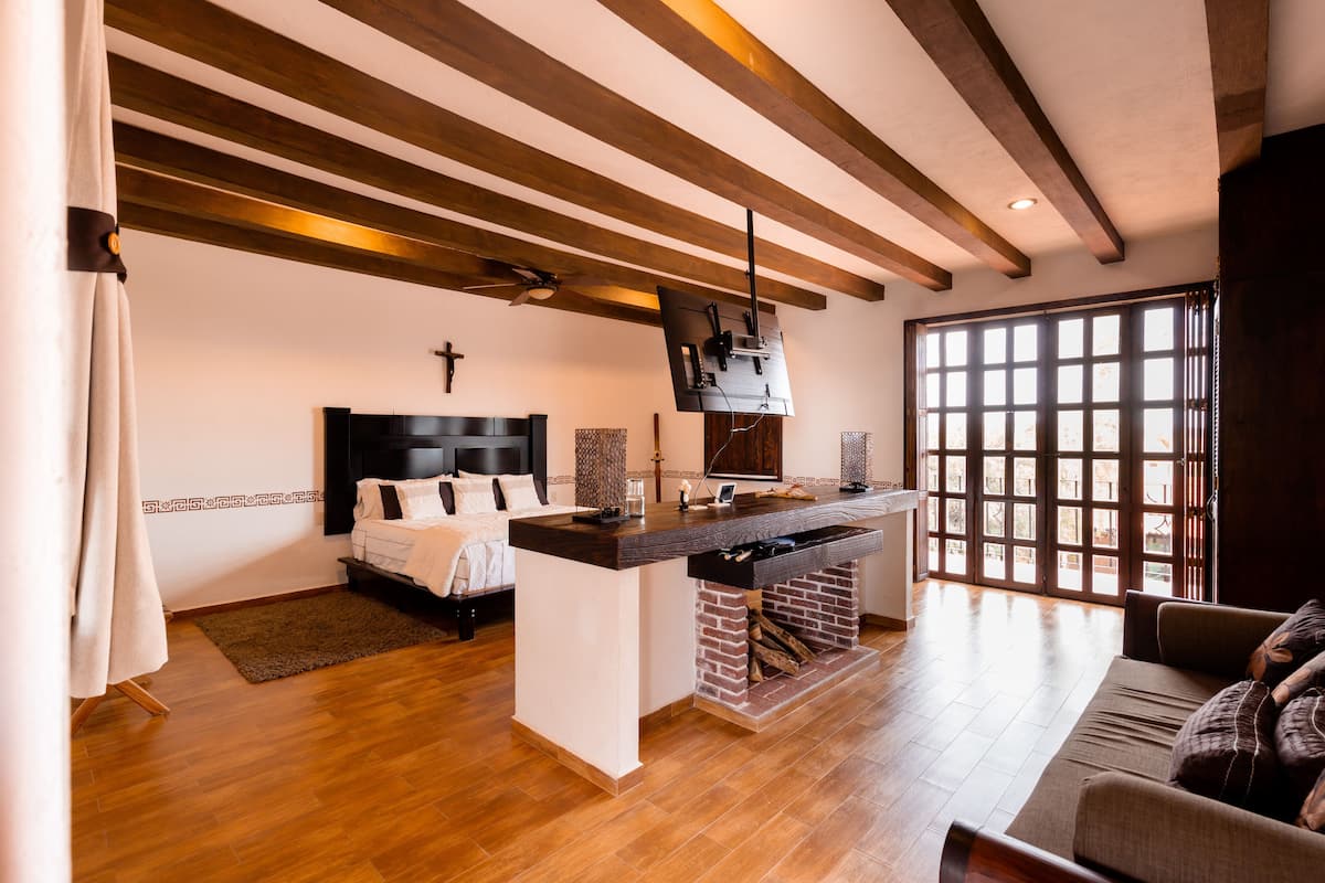 Una habitación de hotel moderna con techo de madera, un ventanal grande, una cama, un sofá y una pequeña cocina.