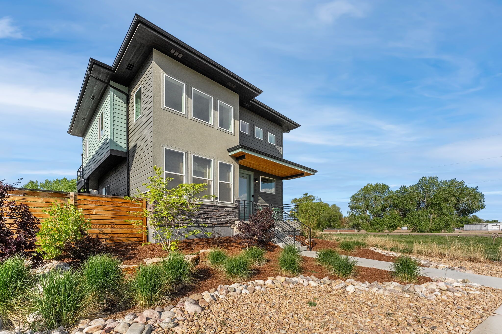 A modern two-story house with a stone facade, a wooden deck, and a landscaped garden with gravel and plants.