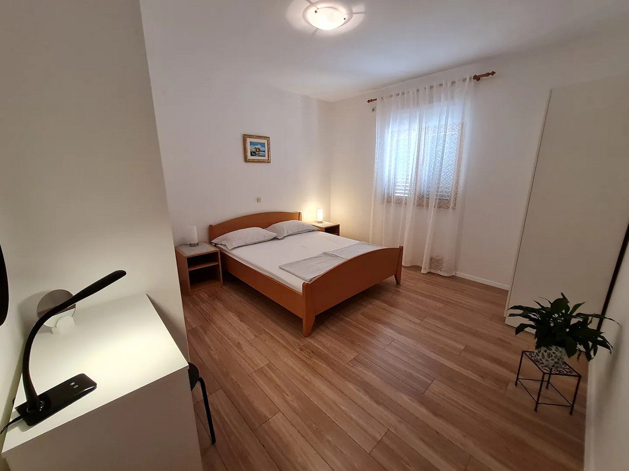 Ein Schlafzimmer mit einem hölzernen Bett, einem Nachttisch mit Lampe, einem kleinen Nachttisch mit Lampe, einer Topfpflanze und einem Bild an der Wand.