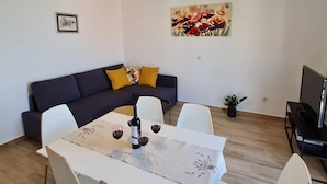 A modern living room with a dark sofa, white chairs, a dining table with a wine bottle and glasses, a painting on the wall, and a TV on a stand. - Apartment Stella (Zupa dubrovacka)