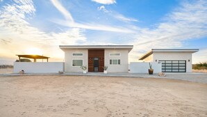 A modern two-story house with a flat roof, a garage, and a covered patio area. - Sun Oro Joshua Tree - Hot Tub, Bbq, Fire Pit & Gameroom 2 Bedroom Home by RedAwning (Joshua Tree)