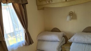 A single bed with a quilted mattress and a headboard, a bedside table with a lamp, and a window with curtains. - Caravan for Rent Coral Beach R2v7 Ingoldmells (Skegness)
