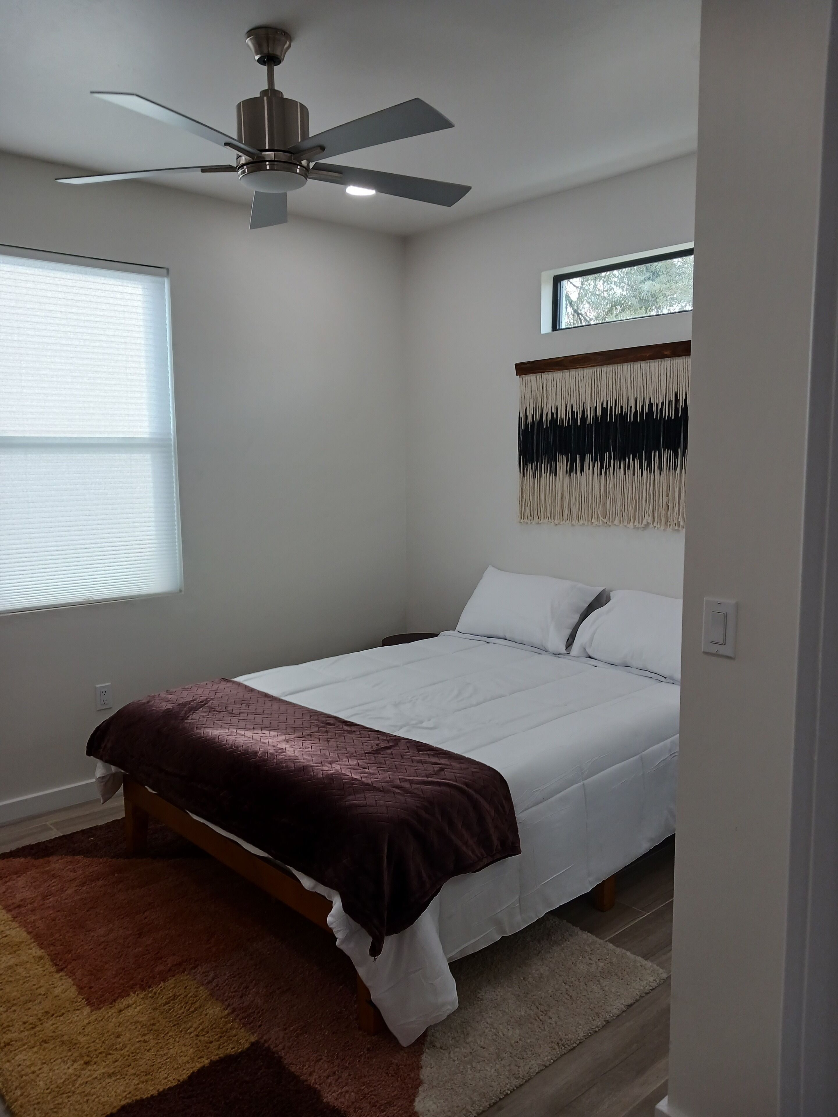 A bedroom with a bed, a ceiling fan, a window, and a wall-mounted macrame piece.