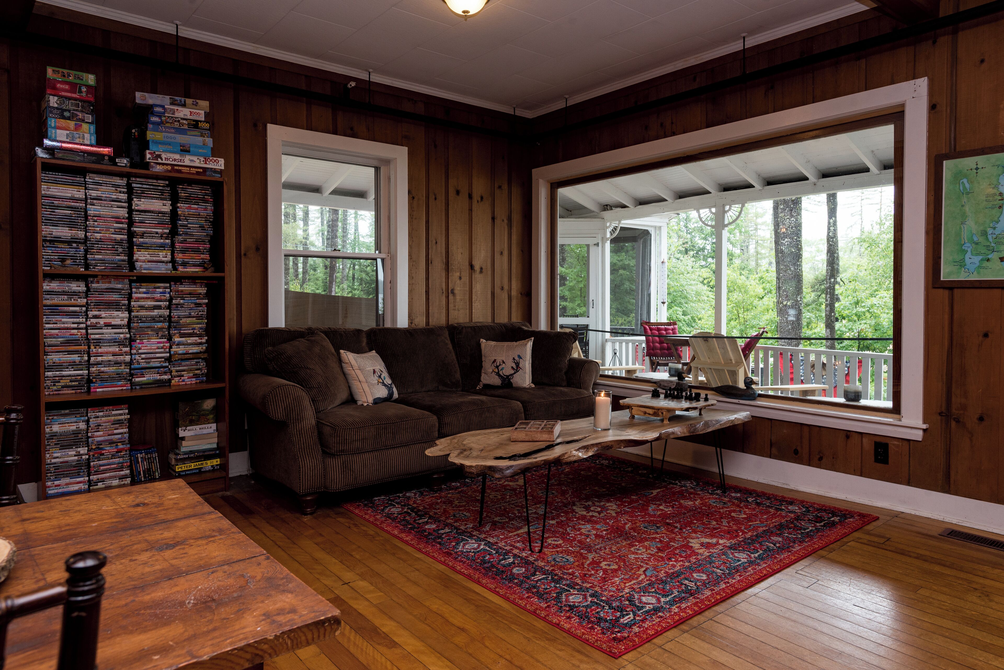 A cozy living room with a wooden floor, a brown leather sofa, a coffee table, and a bookshelf filled with DVDs.
