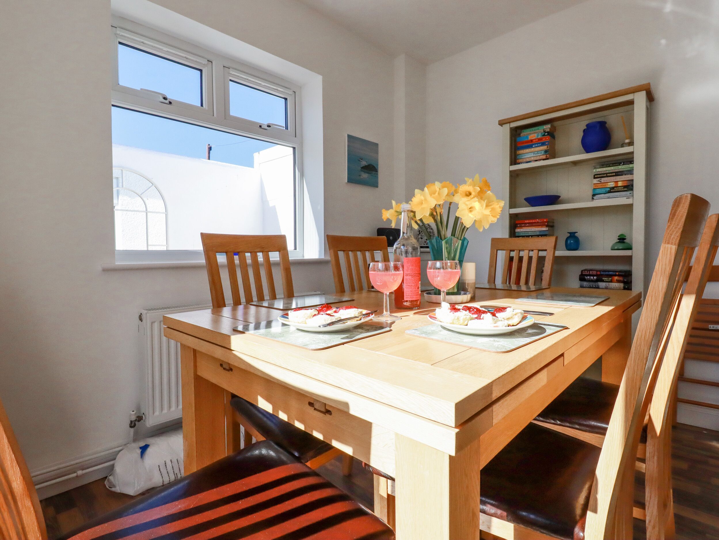 Een eetruimte met een houten tafel, stoelen, een vaas met bloemen en een boekenkast met boeken en decoratieve objecten.