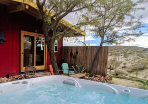 A hot tub area with a wooden deck, a red building, and a view of the desert landscape. - The Cabin Under The Stars - Hot Tub, Bbq And A Firepit 2 Bedroom Cabin by RedAwning (Joshua Tree)