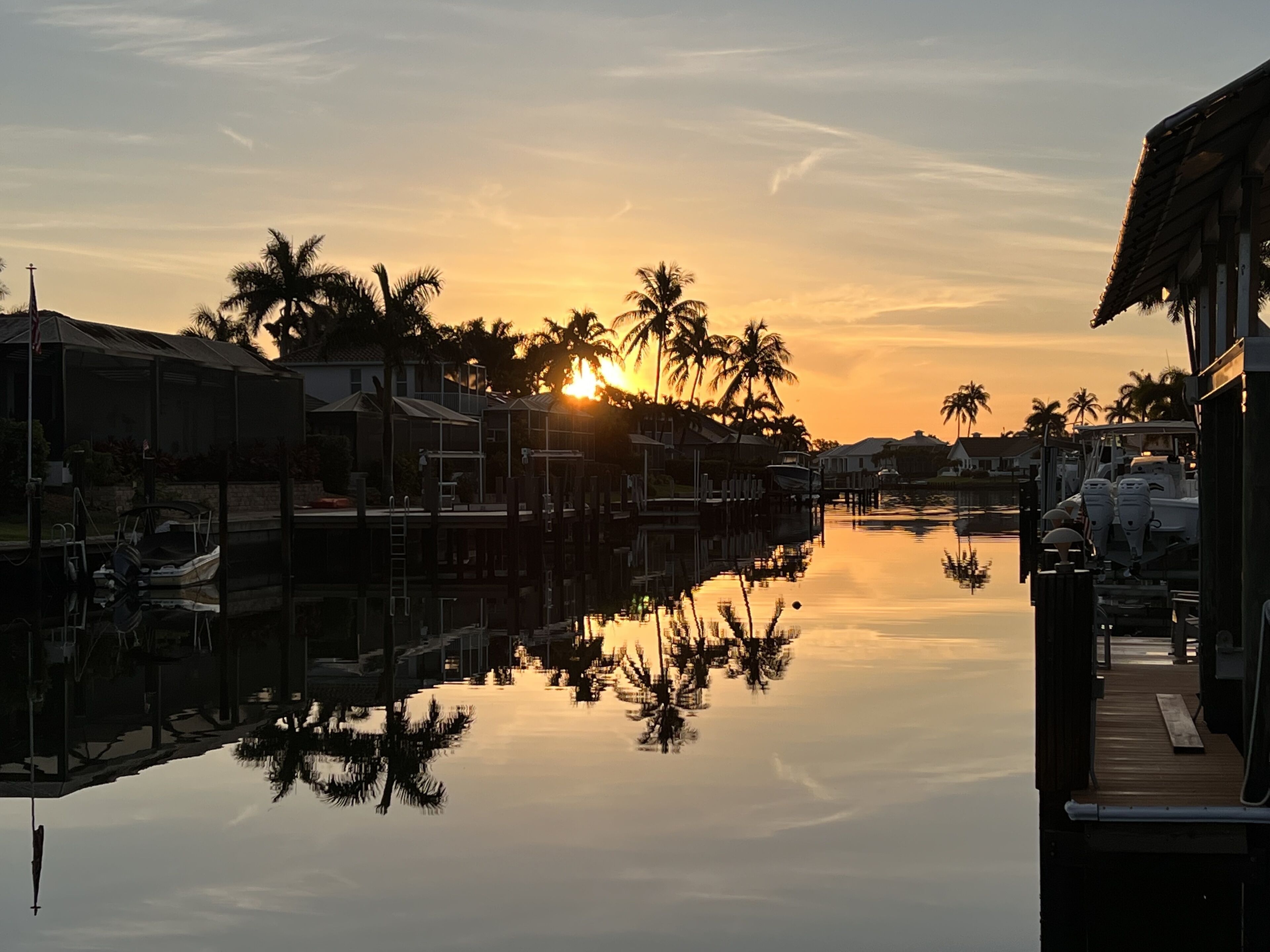 A marina at sunset with calm water reflecting the sky and palm trees.