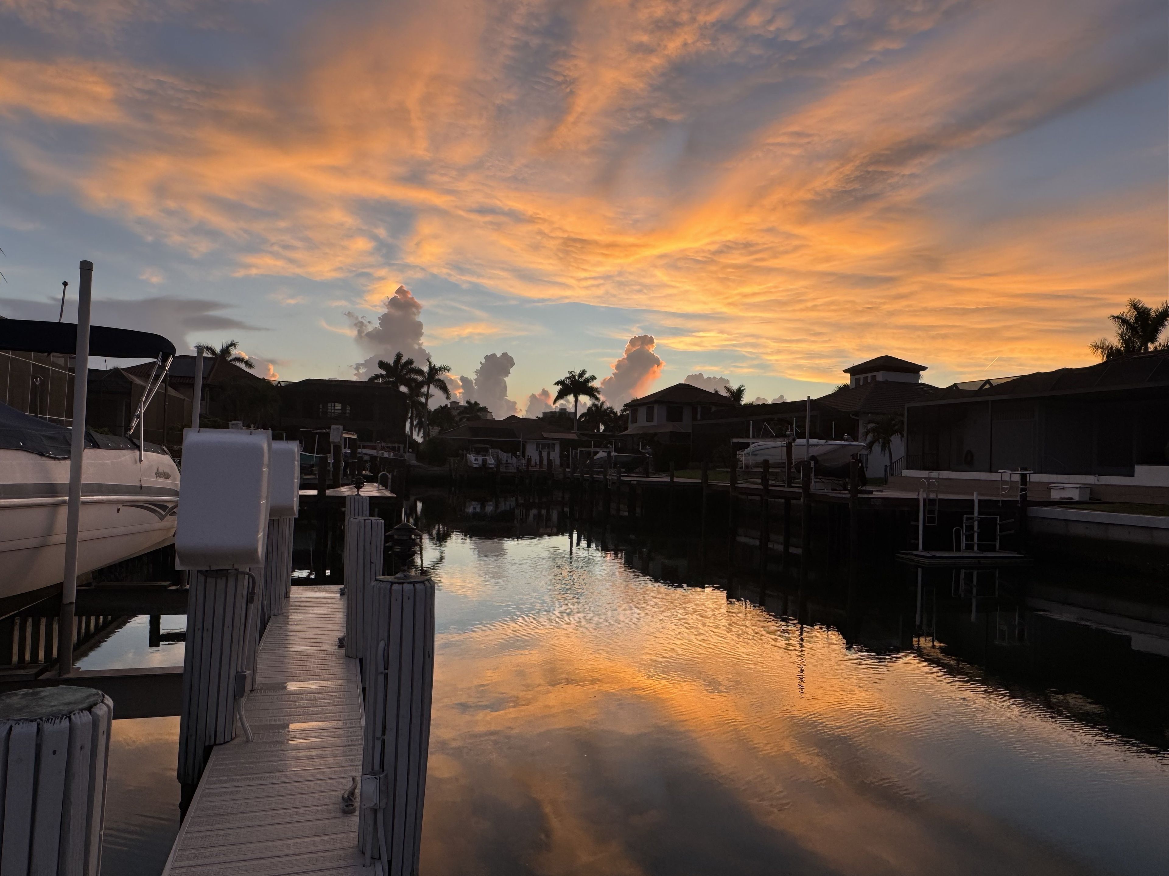 A marina with boats docked, a calm water surface reflecting the sky, and a dock extending into the water.