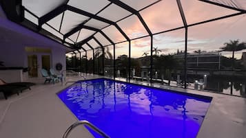 Une piscine intérieure dotée d’un toit en verre, entourée de chaises longues et offrant une vue sur des palmiers et un coucher de soleil.