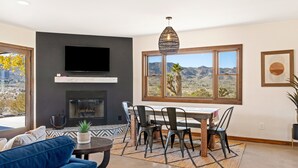 A living room with a TV, fireplace, dining table, chairs, and a view of mountains. - Dreamspace - Hot Tub, Fire Pit, 5 Min. To Park 2 Bedroom Home by RedAwning (Joshua Tree)