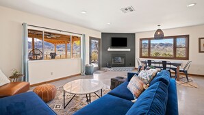 A spacious living room with a large sofa, a round coffee table, and a fireplace. There are large windows offering a view of the mountains. - Dreamspace - Hot Tub, Fire Pit, 5 Min. To Park 2 Bedroom Home by RedAwning (Joshua Tree)
