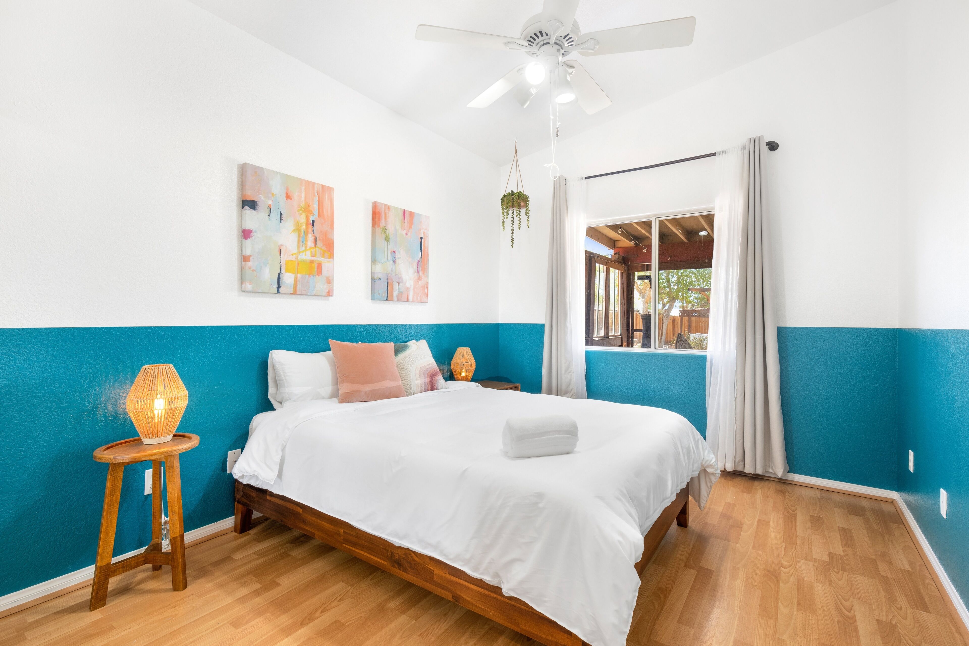 A bedroom with a bed, a ceiling fan, a window with curtains, and artwork on the wall.