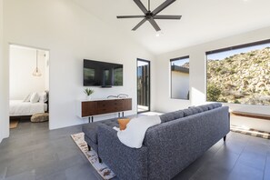 A modern living room with a grey sofa, a flat-screen TV, a ceiling fan, and a large window offering a view of a rocky landscape. - A Little Tish - Secluded Stargazers Getaway 2 Bedroom Home by RedAwning (Yucca Valley)