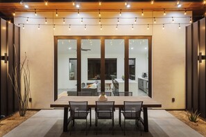 An outdoor dining area with a wooden table and chairs, string lights overhead, and a view into the house through large glass doors. - A Little Tish - Secluded Stargazers Getaway 2 Bedroom Home by RedAwning (Yucca Valley)