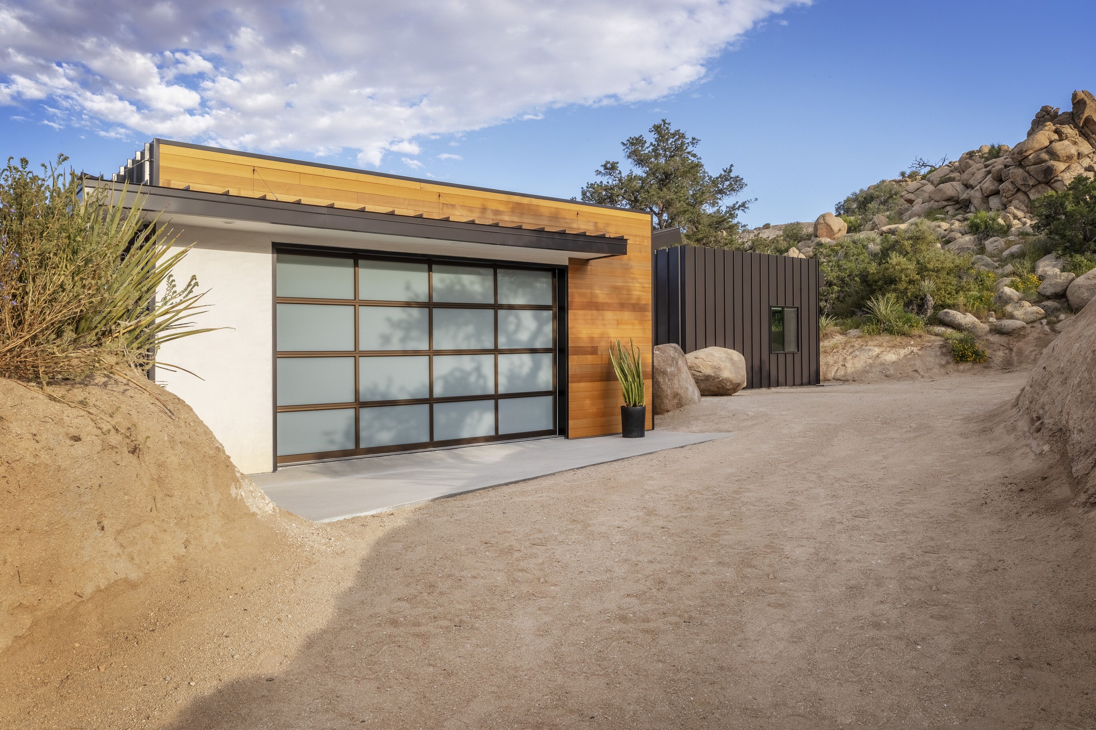 A modern house with a large garage door, a wooden exterior, and a desert landscape with rocks and sparse vegetation.