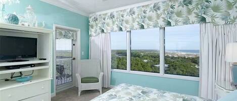 A bedroom with a bed, a television on a stand, a window with curtains, and a view of greenery outside.