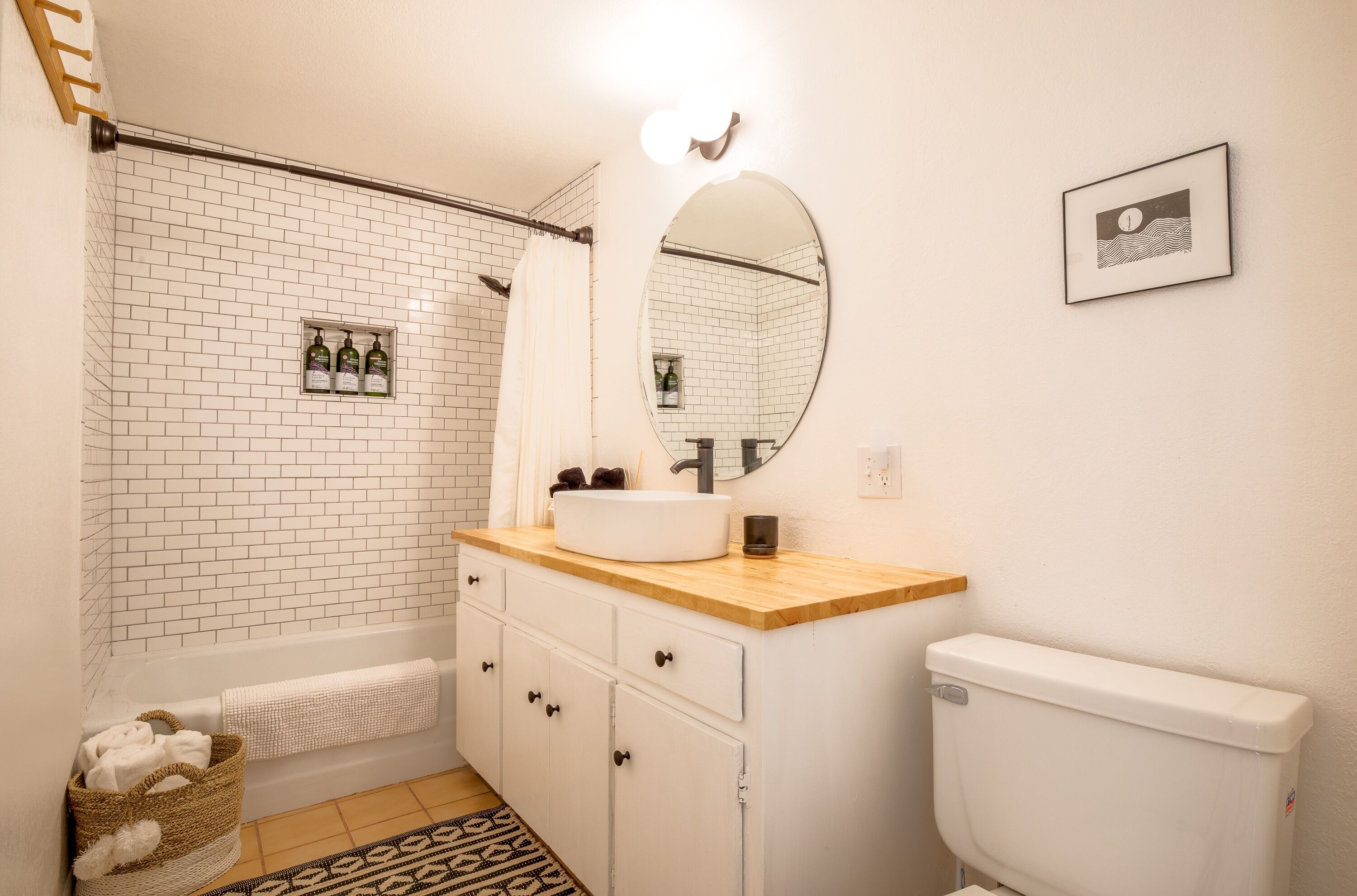 A bathroom with a white tiled wall, a wooden vanity, a white toilet, a mirror, and a framed picture on the wall.