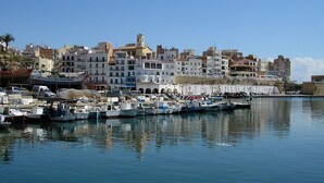 A coastal marina with boats docked, a clear sky, and buildings in the background.