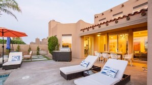 Zona de piscina amb gandules, una taula i cadires, i un edifici amb llum càlida a l'interior.