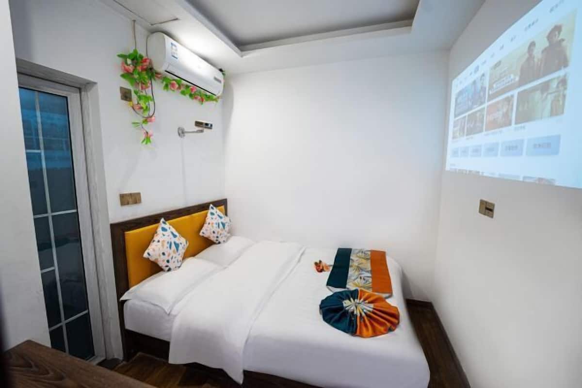 Habitación de hotel con cama, cabecera de madera, aire acondicionado instalado en la pared y una pantalla de proyección que muestra imágenes.