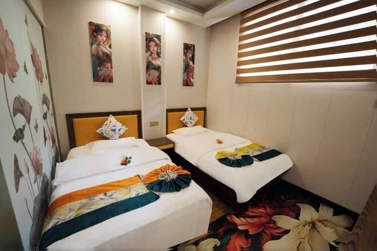 Habitación de hotel con dos camas, cada una con un cabecero decorativo y almohadas. Se observan obras de arte florales en la pared sobre las camas y una ventana con persianas.